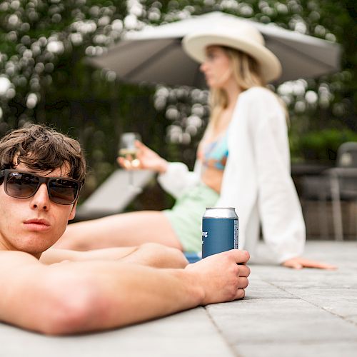 Two people are relaxing by a pool, holding canned drinks. One person is in focus, wearing sunglasses, and the other is blurred in the background under an umbrella.