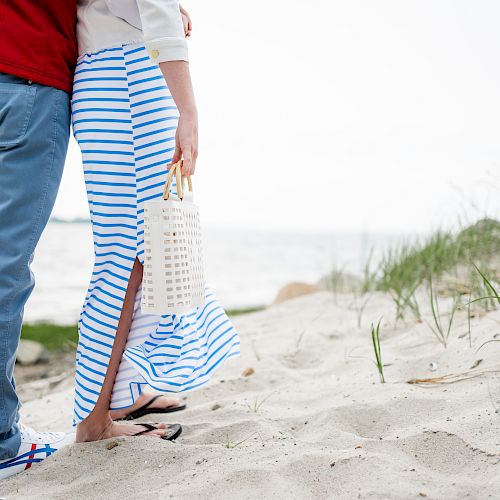 A couple stands on a sandy beach, partially embracing. They wear casual clothes and flip-flops, one holding a striped beach bag, facing the water.