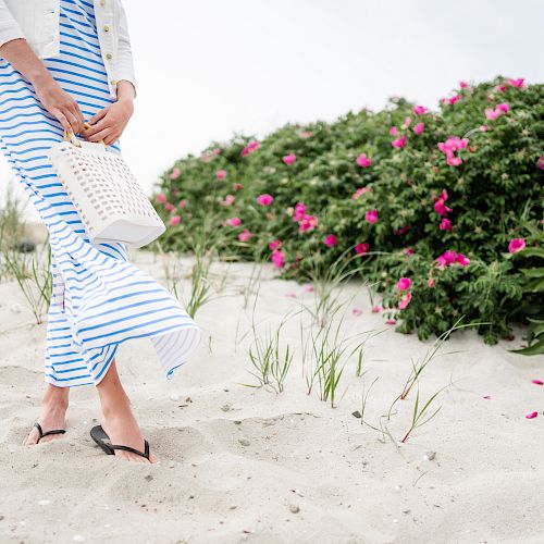 A person in a striped blue and white dress walks on a sandy beach carrying a white basket, with pink flowers and greenery in the background.