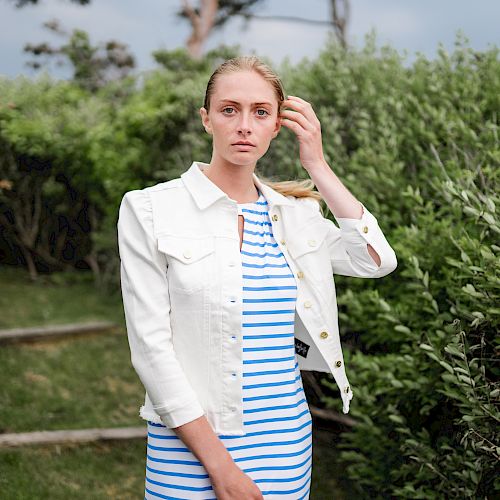 A person wearing a blue and white striped dress with a white jacket stands outdoors, surrounded by greenery, with one hand touching their hair.