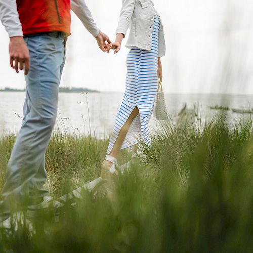 A couple is holding hands while walking through tall grass near a body of water.