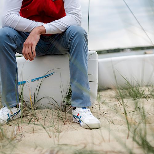 A person is sitting on a white container on a sandy beach, with boats in the background and grass in the foreground.