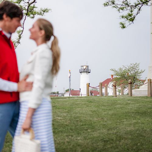 A couple standing closely, facing each other, with a lighthouse, trees, and buildings in the background on a grassy area.