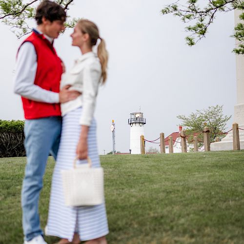 A couple stands on a grassy area near a lighthouse, with a tree overhead. They appear to be in a romantic moment, facing each other closely.