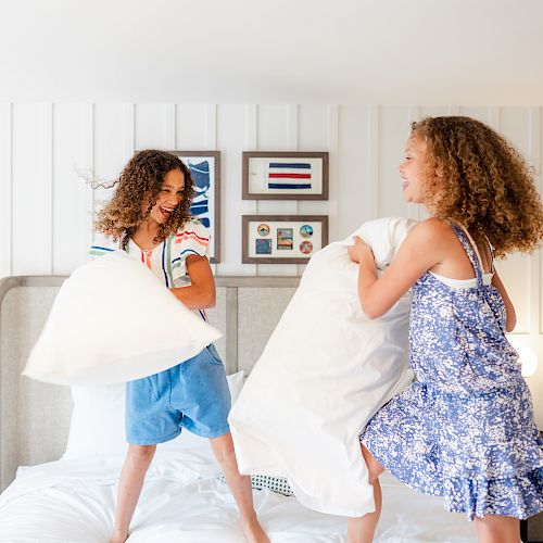 Two children are having a pillow fight on a bed in a brightly lit room with framed pictures on the wall behind them.