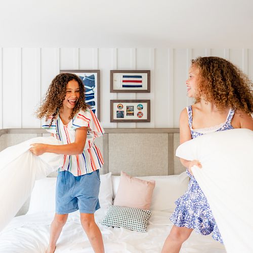 Two kids are having a pillow fight on a bed in a bright room with wall decor and a nightstand lamp.