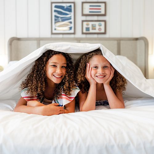 Two children are lying under a white blanket on a bed, smiling and looking at the camera, with framed pictures on the wall behind them.