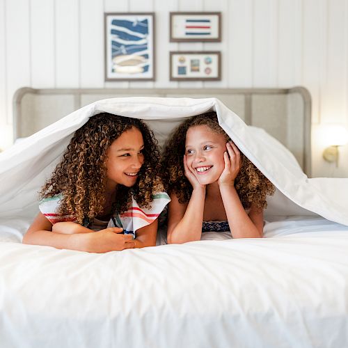 Two children are lying on a bed, partially covered by a white blanket, happily smiling and looking at each other, framed artwork on the wall behind.