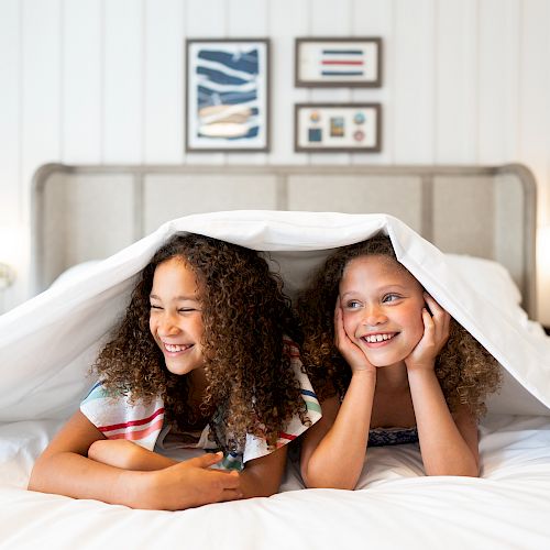 Two children with curly hair lie under a white blanket on a bed, smiling. Artworks are hung on the wall behind them.