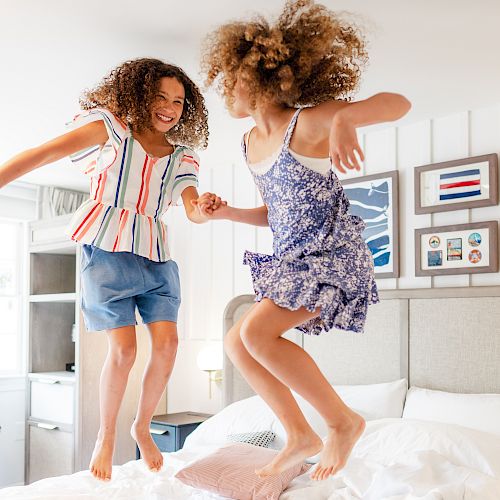 Two children with curly hair are joyfully jumping on a bed in a bright, well-decorated room.