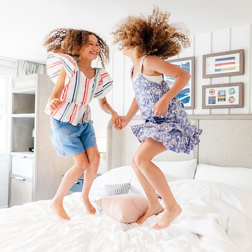 Two smiling children are joyfully jumping on a bed, holding hands, in a bright, well-decorated bedroom.