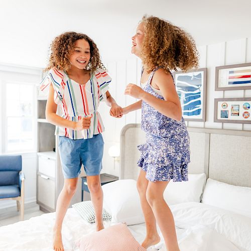 Two children are jumping on a bed, smiling and having fun. The room is decorated with framed pictures and has a bright, airy feel.