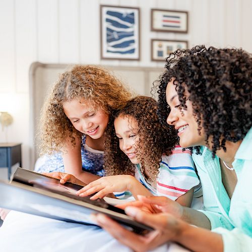 A woman and two children are happily looking at a photo album together while sitting on a bed in a cozy, well-decorated room.