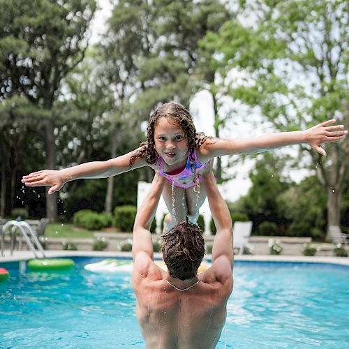 A person lifts a child in a swimming pool, with the child spreading their arms like an airplane and smiling. Trees and poolside chairs are in the background.