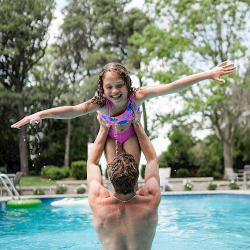 A man is lifting a smiling girl in a swimming pool, both enjoying their time under a bright sky. The girl is spreading her arms like an airplane.