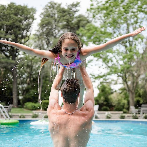 A man is lifting a smiling girl in a pool with outstretched arms, surrounded by trees and pool toys, creating a joyful and fun atmosphere.