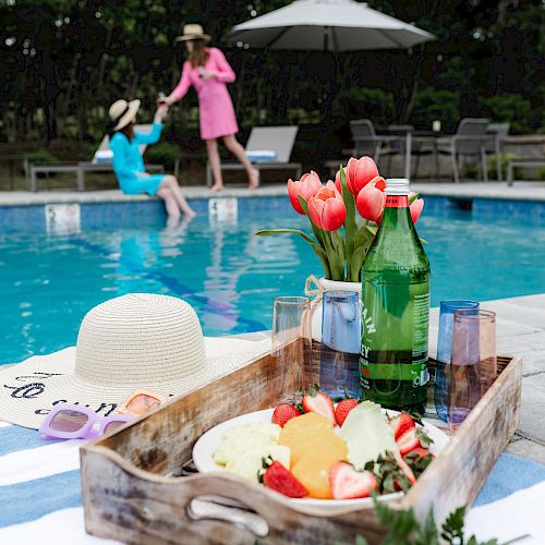 A poolside scene with two people, one in the pool and one standing, plus a tray with food, drinks, a hat, and flowers on a striped towel.