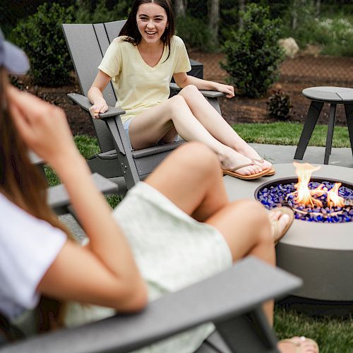 Two people are sitting on outdoor chairs around a fire pit, engaging in conversation in a garden setting.