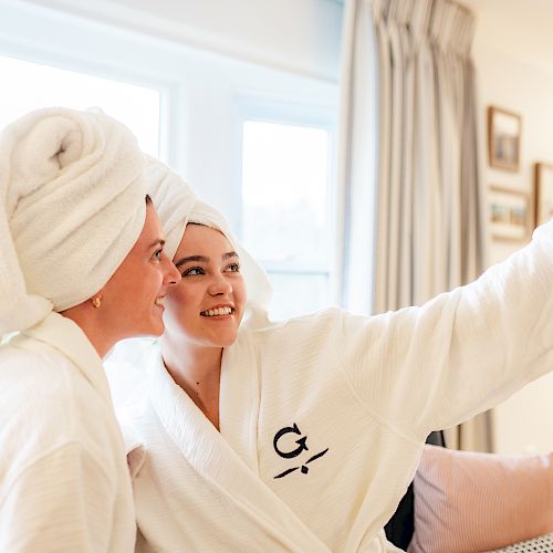 Two people wearing bathrobes and towel wraps on their heads are taking a selfie together in a cozy room.