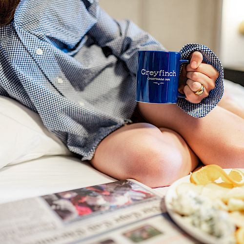 A person in a checkered shirt is sitting on a bed, holding a "Greyfinch" mug, with a plate of food and a newspaper lying nearby.