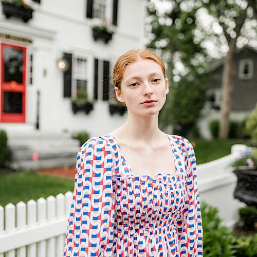 A person in a patterned dress stands in front of a white house with black shutters and a red door, alongside a white picket fence and greenery.