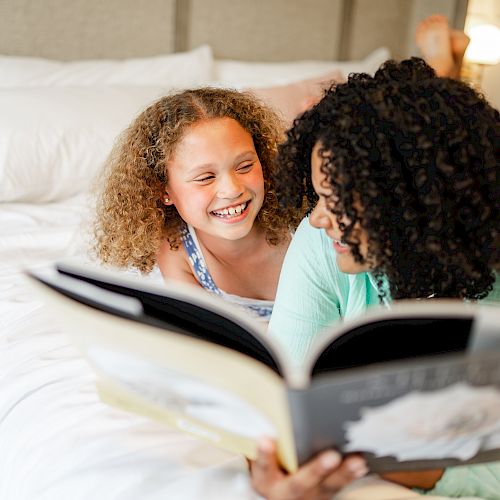 A person and a child are lying on a bed, smiling and engaging with a large book together, creating a joyful and relaxed atmosphere.