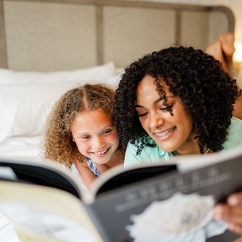 A woman and a child are lying on a bed, smiling and reading a book together. The background has framed pictures on the wall. Ending the sentence.