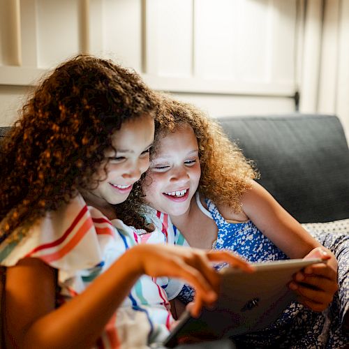 Two children are sitting on a couch, smiling and looking at a tablet together, with a lamp illuminating the scene.