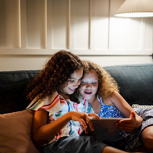 Two children are seated on a couch under a lamp, smiling and looking at a tablet screen while sitting close to each other.