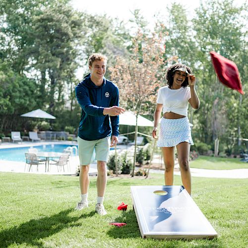 Two people are playing cornhole near a pool on a grassy lawn. One throws a red beanbag while the other watches and cheers.