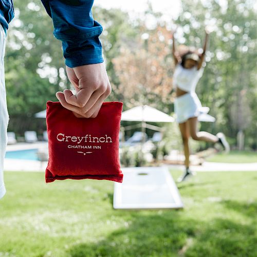 A person holds a red Greyfinch bean bag while another person leaps into the air near a cornhole board on the grassy yard.