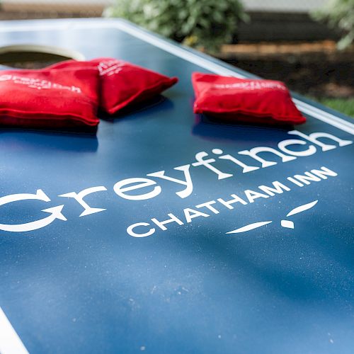 The image shows a blue cornhole board labeled "Greyfinch Chatham Inn" with three red bean bags on top.