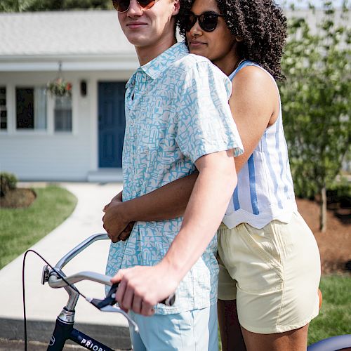 A couple wearing sunglasses stands close together behind a bicycle in front of a house with a blue door, enjoying a sunny day.