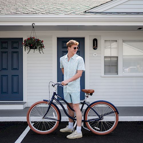 A person stands next to a bicycle in front of a light-colored building with blue doors and windows, wearing casual summer clothing and sunglasses.