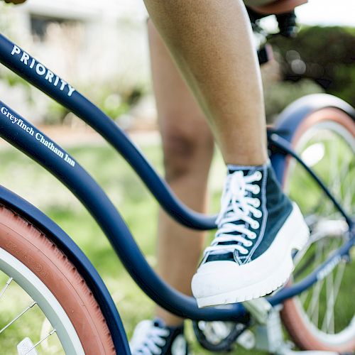 A person wearing black and white sneakers is riding a "Priority" bicycle with a blue frame and red tires. The surroundings are grassy.