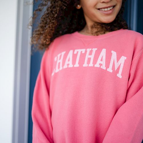 A person wearing a pink sweatshirt with "CHATHAM" written on it, standing in front of a blue door and white siding wall, is smiling.