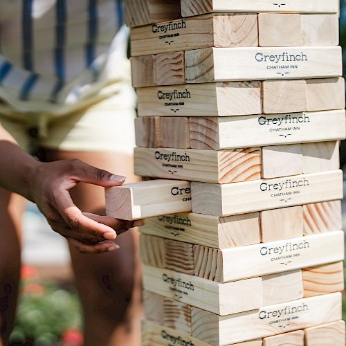 A person is playing a giant outdoor Jenga game with wooden blocks labeled "Greyfinch." The game is set up outside in a garden setting.