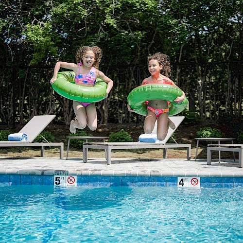 Two kids, each with a green float ring, jump into a swimming pool from the edge with lounge chairs and umbrellas in the background.