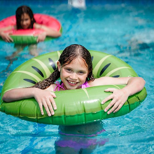 Two children are in a pool, each using an inflatable swim ring. The child in front has a green ring, while the other has a red one, both smiling.