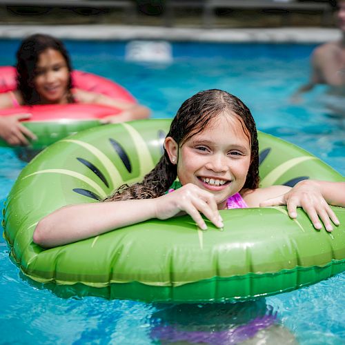 Two kids are in a pool with inflatable rings and are smiling. An adult is in the water behind them, enjoying the sunny day and pool time.