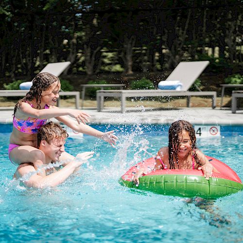 A man carrying a child on his shoulders splashes water at another child on a float in a pool. Sun loungers are visible in the background.