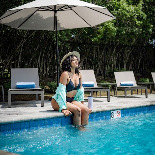 A person in swimwear sits by a pool under a large umbrella with lounge chairs in the background, surrounded by greenery.