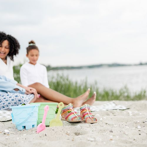 Three people sit on a blanket by the beach, with sand toys and sandals nearby, enjoying their time together by the water.