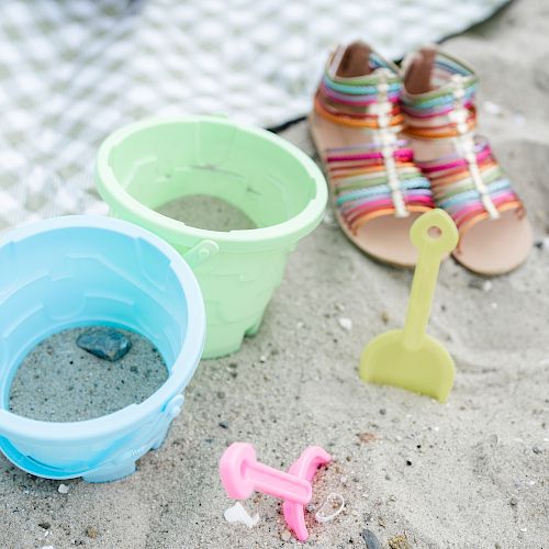 The image shows two sand buckets, a small shovel, and a pair of colorful sandals on the sand near a checkered blanket.