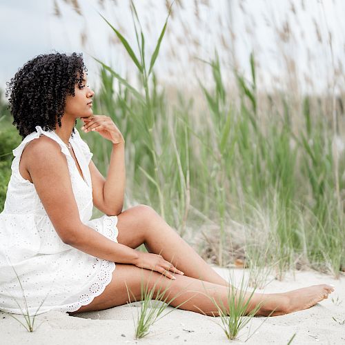 A person in a white dress sits thoughtfully on a sandy beach near tall grass and pink flowers, creating a peaceful and serene setting.