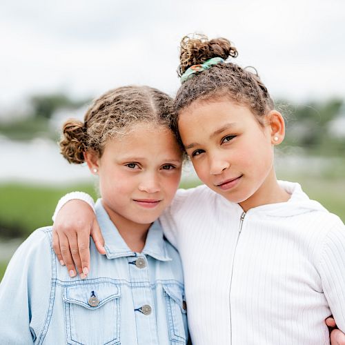 Two young girls stand close together in a grassy area near water, smiling with arms around each other, creating a heartwarming scene.