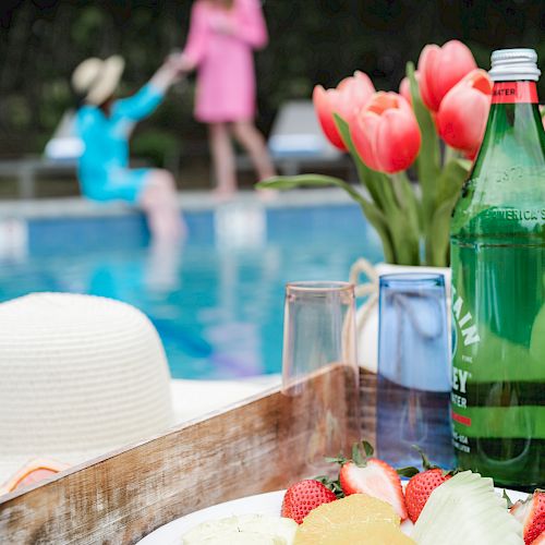 A tray with fruits, a bottle, and glasses by a poolside setting; two people are near the pool, with flowers and a hat in the foreground.