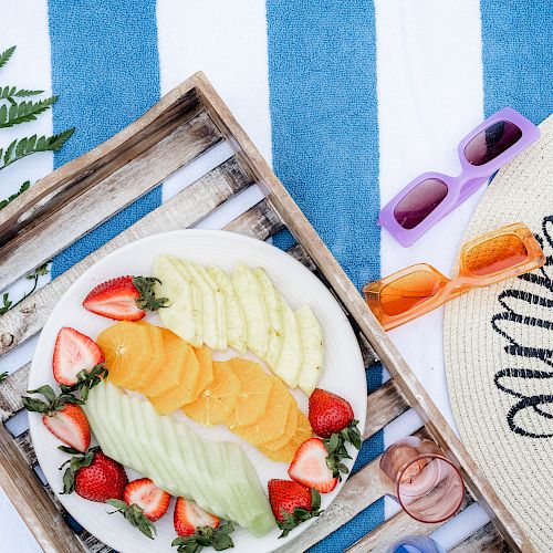 A wooden tray with sliced fruits, sunglasses, a sunhat, and a potted plant on a blue and white striped fabric.