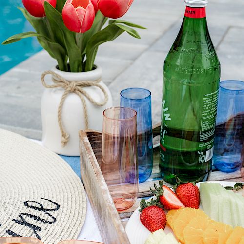 A poolside setting with a hat, sunglasses, a bottle of water, glasses, a vase of red tulips, and a plate of sliced fruits and strawberries.