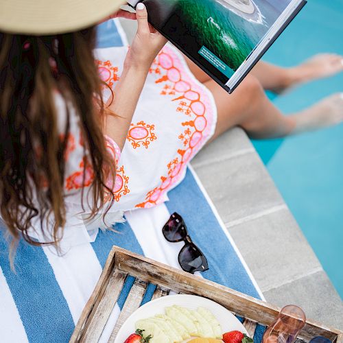 A person is sitting by the pool, reading a book. There is a basket with cut fruit and a bottle of water next to them.